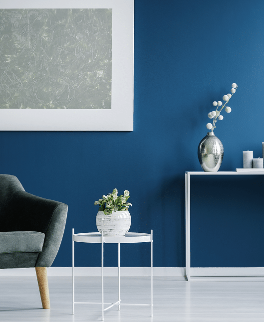 Salon moderne: murs bleu marine. Fauteuil gris, table blanche avec plante. Tableau abstrait argenté encadré. Console blanche avec vase et bougies.