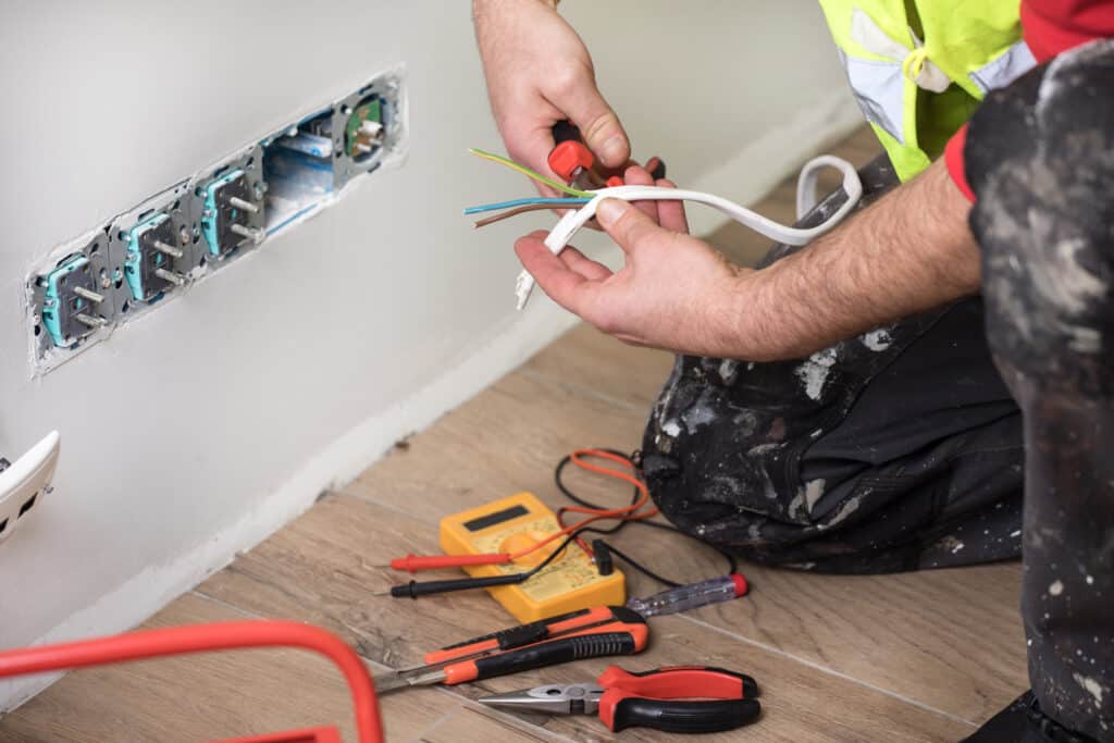 Électricien dénudant des fils colorés (jaune, vert, bleu, marron) pour l'installation de prises murales avec outils sur un sol en bois.