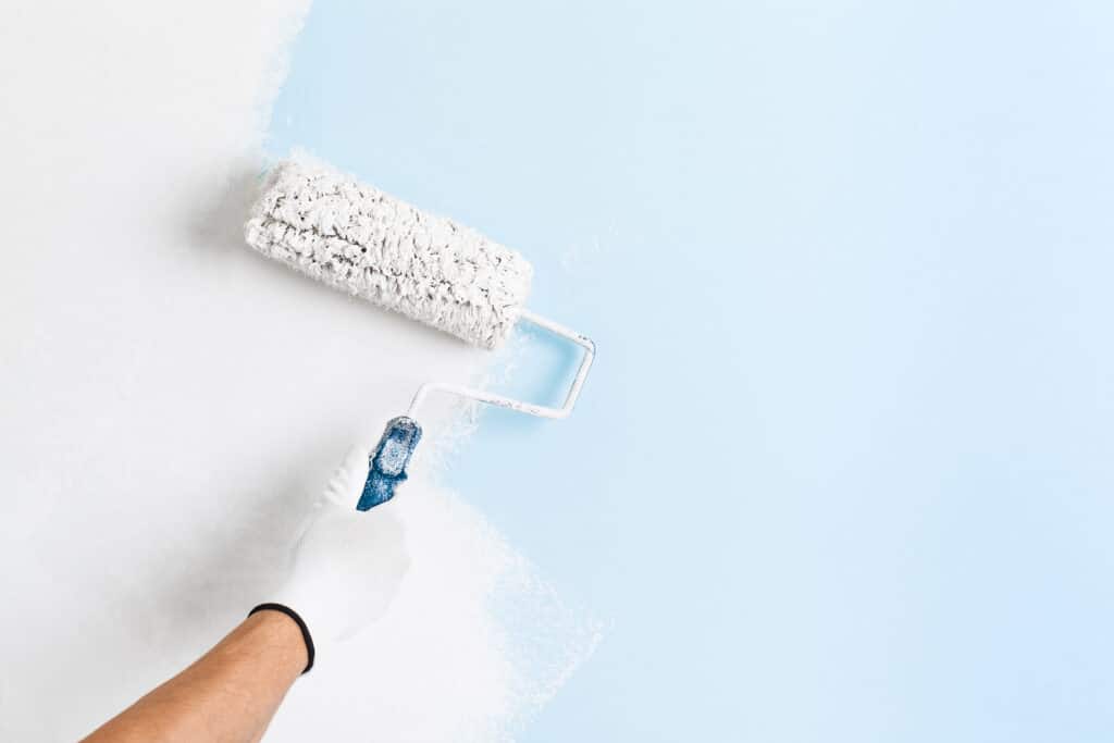 Main gantée appliquant de la peinture blanche avec un rouleau sur un mur bleu clair. Rénovation.