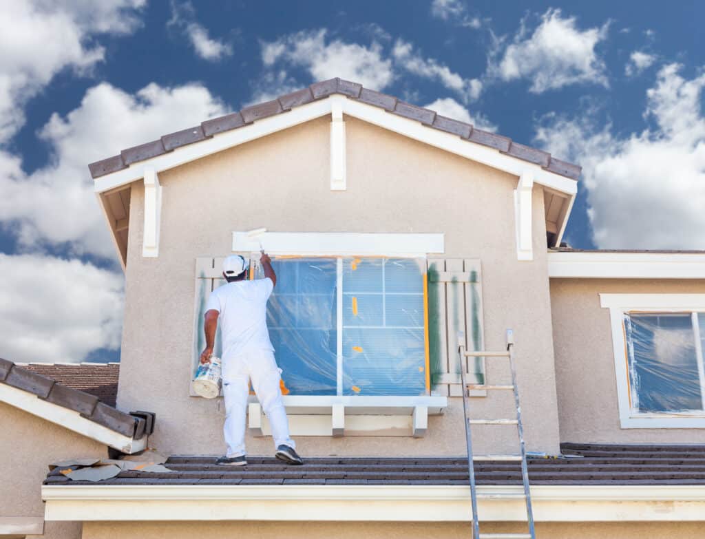 Peintre en blanc sur un toit de tuiles, appliquant de la peinture blanche sur les boiseries d'une fenêtre protégée sur une façade beige. Rénovation de maison.