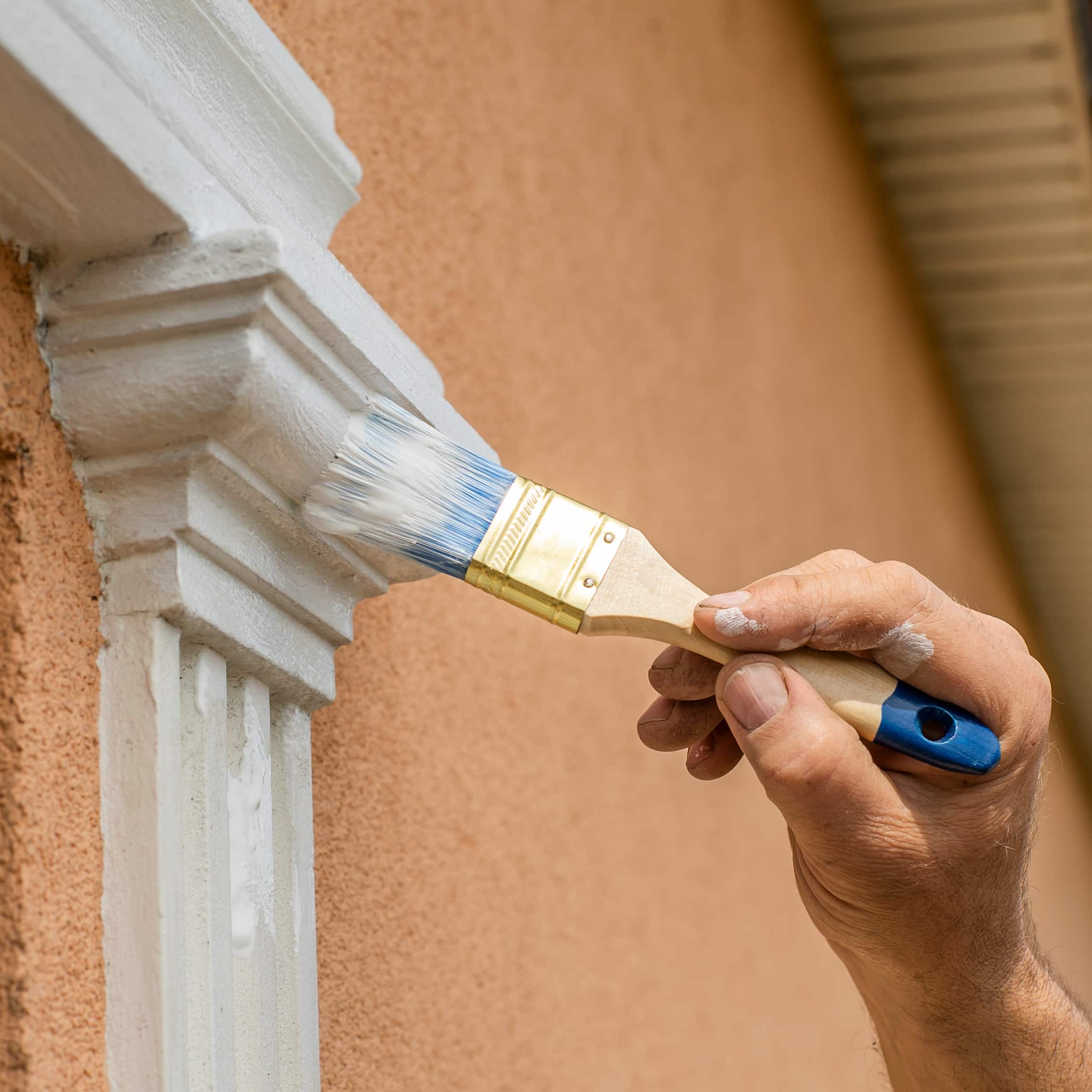 Une main peignant une moulure décorative blanche sur un mur crépi orange avec un pinceau propre, des traces de peinture sur les doigts.