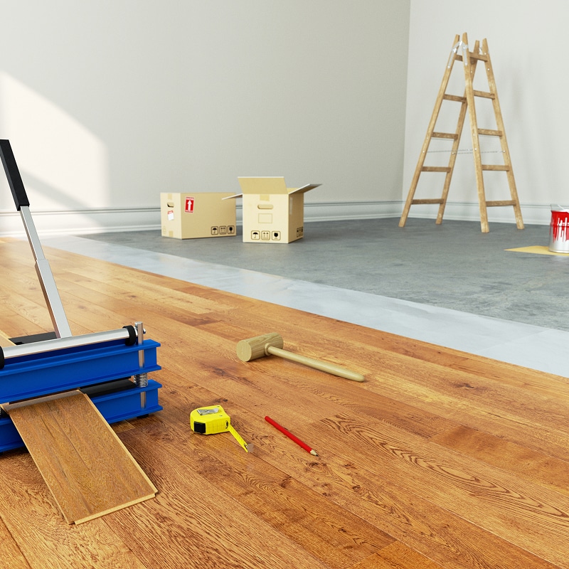 Installation de parquet stratifié brun avec un cutter, maillet, mètre et crayon au sol. Une pièce en rénovation avec cartons et échelle en arrière-plan.