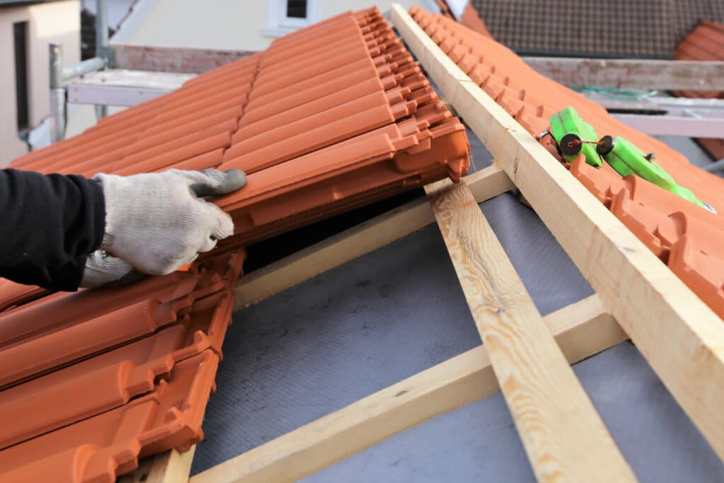 Main gantée d'un artisan posant des tuiles terre cuite sur une toiture neuve. Lattes de bois et sous-toiture gris visible. Chantier construction.